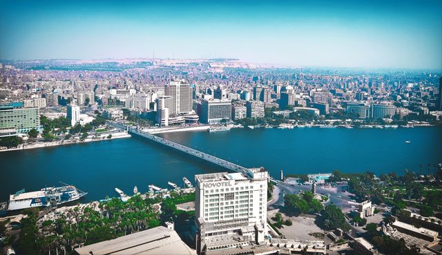 This image depicts an expansive aerial view of Cairo, Egypt, with the river, city buildings, and vibrant urban landscape. The photo can be used in travel brochures, blog posts highlighting cityscapes, articles about Cairo, and promotional materials for tourism.