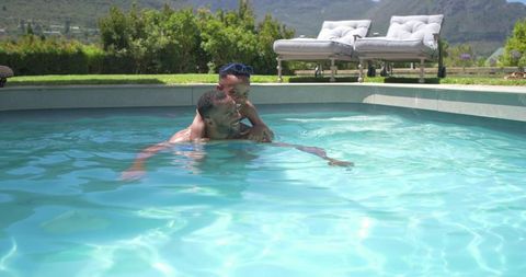 African american father and son playing, laughing in backyard pool on sunny day with sunglasses