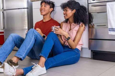 Young couple sitting on kitchen floor syncing smartwatches and sharing morning coffee