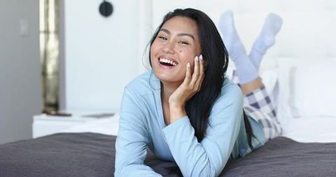 Asian woman laughing on bed in cozy loungewear, relaxing morning at home