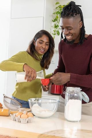 Diverse couple baking together in bright modern kitchen pouring milk and sifting flour