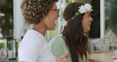 Friends sharing laughter at garden brunch on sunroom patio, floral crown, pastel dresses