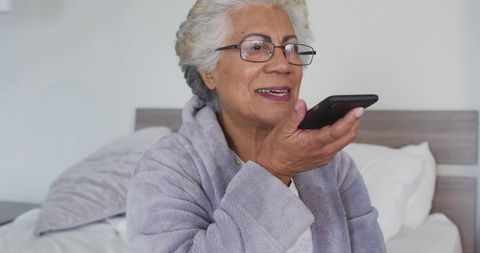 Senior woman speaking into smartphone while relaxing in bathrobe on bedroom bed