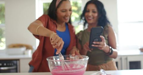 Diverse friends whisking pink batter at sunlit kitchen island following recipe on phone