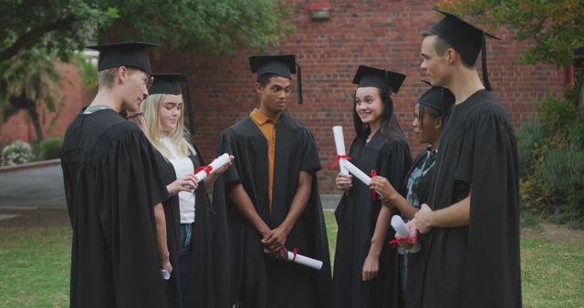 Shows a group of diverse students celebrating graduation outdoors in academic gowns with diplomas in hand. Perfect for use in education, success, achievement, college promotional material, university brochures, student life blogs, and articles about diversity in education.