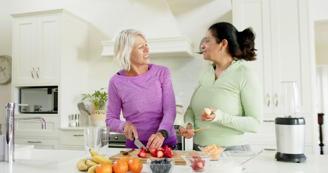 Two women preparing a meal in a modern kitchen setting, cutting fresh fruits and vegetables while smiling and talking. They wear casual clothing appropriate for light exercise or leisure. This can be used in contexts promoting healthy eating, friendship, lifestyle, and kitchen activities.