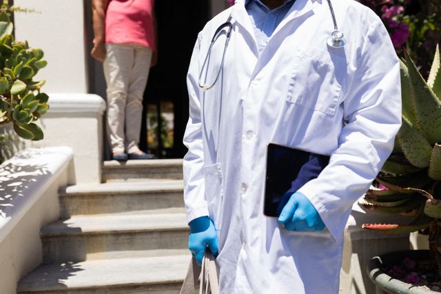 Senior African American male doctor standing outside holding a digital tablet, wearing gloves and a stethoscope. An African American woman is in the background. This image can be used for articles on healthcare, elderly care, COVID-19 pandemic, and retirement lifestyle.