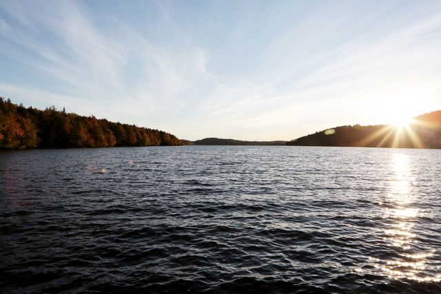 This stock photo depicts a tranquil scene of a calm lake surrounded by trees in autumn colors during sunset. The light reflecting on the water enhances the serenity of the landscape. Suitable for use in promotions related to nature, tranquility, and outdoor activities. Ideal for blog posts, website headers, and travel brochures aimed at showcasing peaceful environments.