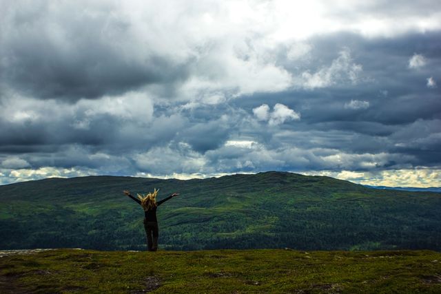 Person standing on a hilltop with arms raised, overlooking a vast green mountain range with dramatic, cloudy sky. Perfect for depicting wandering spirit, outdoor adventure, exploration, and freedom. Ideal for travel blogs, nature documentaries, adventure magazines, and promoting outdoor activities.