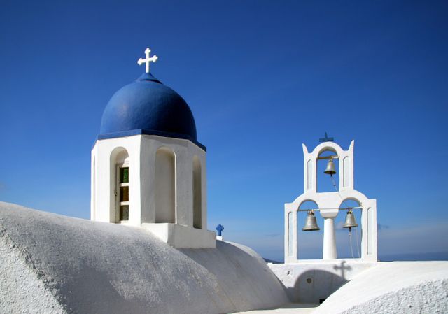 White church with blue dome and bell tower against clear blue sky. Suitable for use in travel brochures, websites promoting Greek tourism, Mediterranean culture magazines, or to illustrate articles about Greek architecture. Ideal for content about Santorini, holidays, and traditional Cycladic architecture.