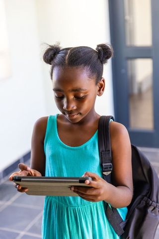 African american schoolgirl using tablet with backpack in bright sunlit school corridor