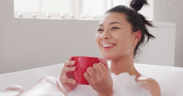 Image of portrait of smiling biracial woman sitting in bathtub in bubble bath with red cup of tea. Health and beauty, leisure time, domestic life and lifestyle concept.