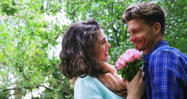 A Caucasian couple enjoys a romantic moment outdoors, with the woman holding a bouquet of pink flowers, with copy space. Their affectionate embrace and joyful expressions convey a sense of love and happiness in a natural setting.