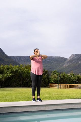 Mature woman practicing mindful standing pose by pool with mountain backdrop and lawn