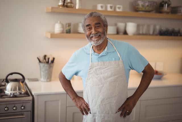 Senior man standing in a modern kitchen, smiling with hands on hips. He is wearing a light blue shirt and an apron, suggesting he is cooking or preparing food. The background includes kitchen shelves with bowls and glasses, a kettle on the stove, and various utensils. This image can be used for themes related to home cooking, senior lifestyle, happiness, and domestic activities.
