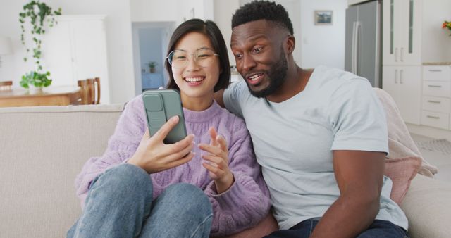 Diverse couple sitting on couch and using smartphone in living room. Spending quality time at home concept.