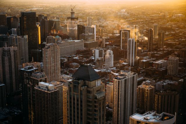 This image captures an aerial view of an urban downtown cityscape during sunset, highlighting high-rise buildings and skyscrapers bathed in a golden hue. Ideal for presentations about urban development, architecture, real estate, and city planning. Suitable for use in travel blogs, websites about city living, and promotional materials for businesses or events located in metropolitan areas.