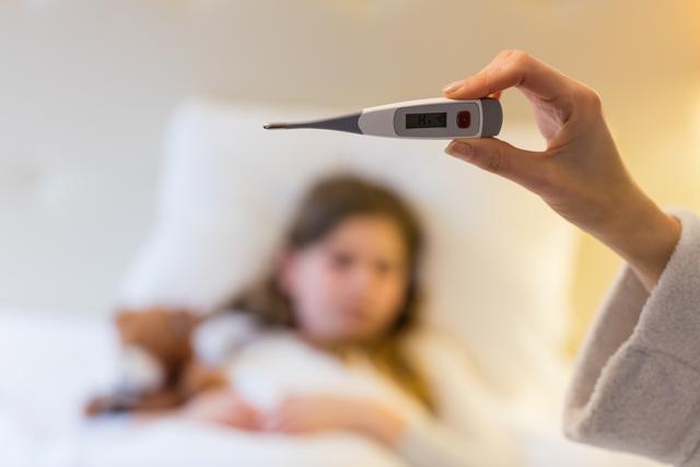 Mother holding digital thermometer while checking child's temperature in bedroom. Child lying in bed, appearing unwell. Useful for topics on family healthcare, parenting, illness recovery, home medical care, and monitoring fever.