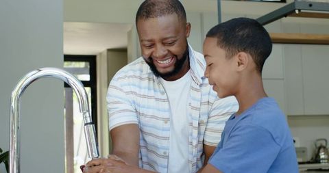 African american father teaching son hand washing at kitchen sink, joyful parenting moment