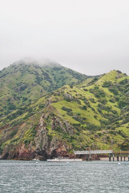 Tranquil coastal scenery featuring green hills with fog gently covering the mountain top. The calm water and a pier combine to create a serene atmosphere. Ideal for travel websites, nature blogs, promotional materials conveying peace, adventure, and beautiful nature views.