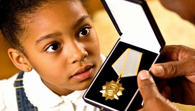 Young girl gazing at gold star medal in presentation box while hands offering award