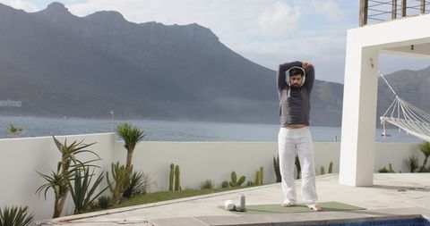 Indian man stretching on coastal terrace yoga mat overlooking sea and mountains