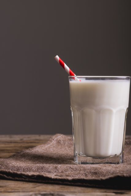 This image shows a close-up of a glass of milk with a red striped straw placed on a napkin on a wooden table against a gray background. Ideal for use in articles about healthy eating, dairy products, nutrition, or rustic kitchen settings. Perfect for food blogs, recipe websites, or advertisements for dairy products.