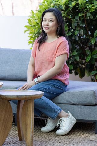 Asian woman sitting on patio sofa in coral blouse and jeans wearing smartwatch