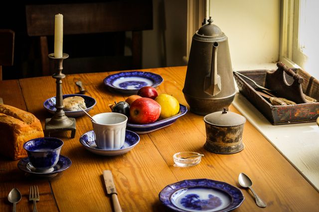 Rustic table set up for breakfast with fresh fruits and bread. Ceramic plates, cups, and vintage items including a candlestick and coffee pot are placed neatly. Ideal for content focusing on rustic or farmhouse style living, morning routines, cozy kitchens, food preparation, family gatherings, and vintage aesthetics.