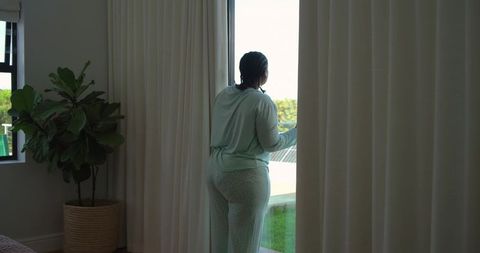 Mid-adult african american woman standing at balcony door looking outside in mint pajamas