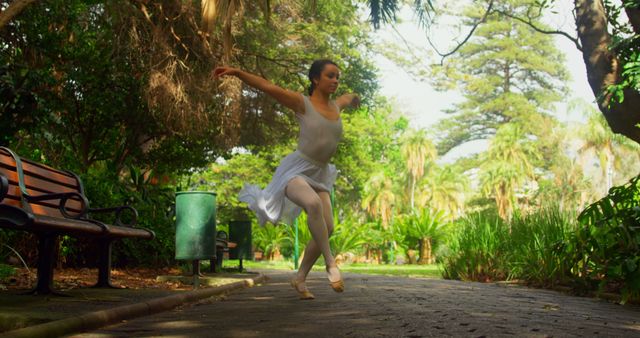 Ballerina dressed in traditional ballet attire performing dance moves surrounded by lush greenery in park on a sunny day. Useful for themes related to dance, nature, performance art, fitness, and outdoor activities.