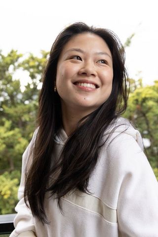 Asian woman smiling on balcony wearing light hoodie and gold earrings, lush greenery