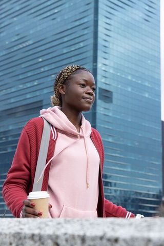 African american woman walking urban streets holding coffee in pink hoodie with backpack