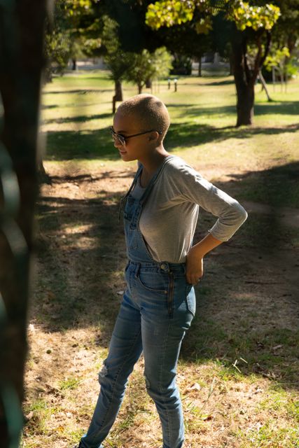 Biracial woman with short blonde hair and sunglasses is walking in a park on a sunny day, wearing denim dungarees and a casual outfit. This image is perfect for use in lifestyle blogs, fashion articles, urban living promotions, and advertisements focusing on independent and modern women enjoying outdoor activities.