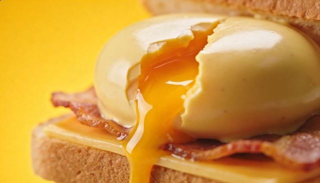 Golden runny egg yolk oozing over bacon and cheese on toasted bread, vibrant brunch close-up