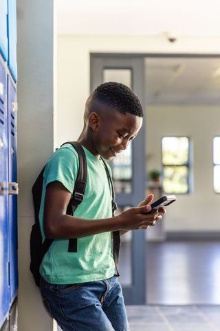 African american boy leaning on locker smiling while using smartphone, backpack at school