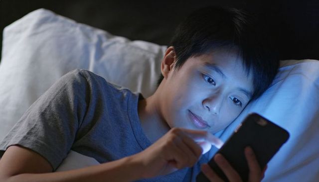 Teen boy lying in bed using smartphone at night blue screen glow closeup