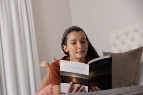 Woman reading sunset photo book while lying on bed in cozy neutral bedroom