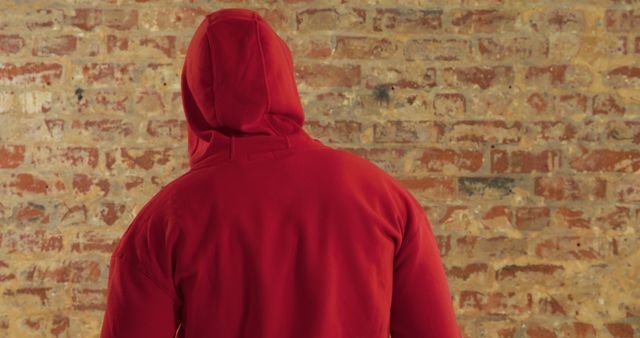 Caucasian man in red hoodie stands facing a brick wall. His identity is obscured, adding a sense of mystery or anonymity to the scene.