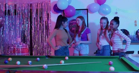 Diverse female friends chatting around pool table at neon birthday party with balloons