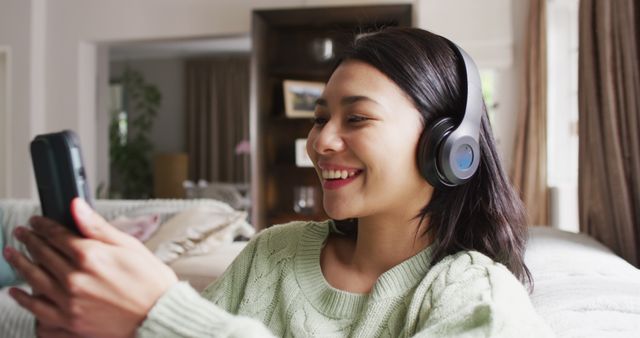 Image of smiling biracial woman with dark hair using headphones and smartphone. Leisure time, domestic life and wellbeing lifestyle concept.