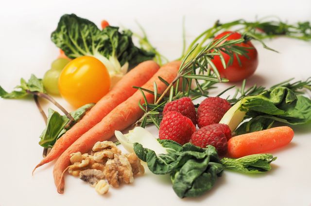This stock photo shows a variety of fresh, organic vegetables and fruits spread out on a white surface. The colorful display includes carrots, yellow and red tomatoes, raspberries, walnuts, and various leafy greens, illustrating a healthy and diverse diet. This image can be used for articles, blog posts, and marketing materials related to nutrition, healthy eating, vegan diets, and organic produce.