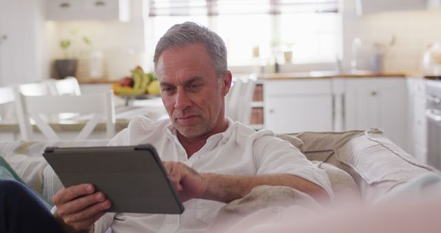 Middle-aged man with grey hair sitting comfortably on sofa in bright, modern kitchen using tablet. Suitable for use in articles about technology usage among older adults, digital lifestyle, and home settings.