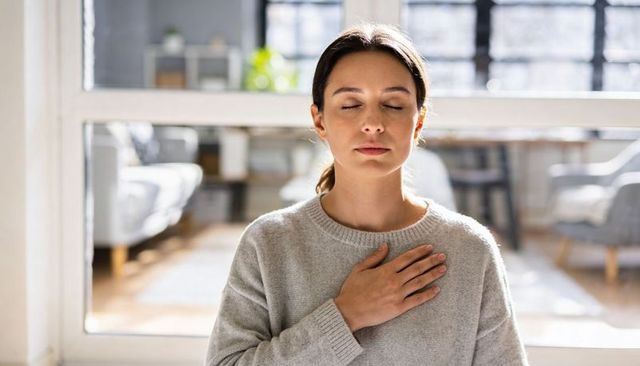 Young woman practicing mindful deep breathing at home hand on heart calm interior