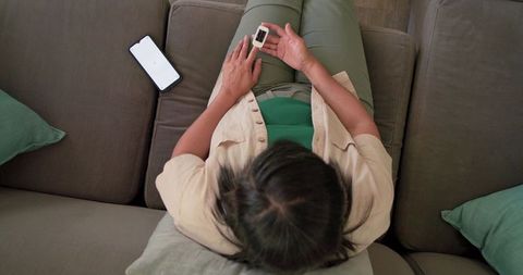 Mature asian woman checking wearable oximeter while relaxing on couch with smartphone