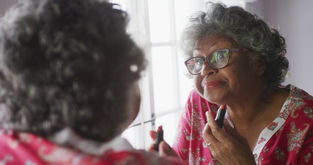 Senior biracial woman applies lipstick at home, with copy space. She's reflected in a mirror, adding a touch of elegance to her morning routine.