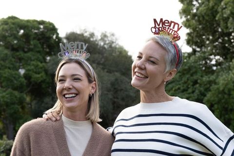 Two women wearing merry christmas headbands smiling together outdoors in lush green park