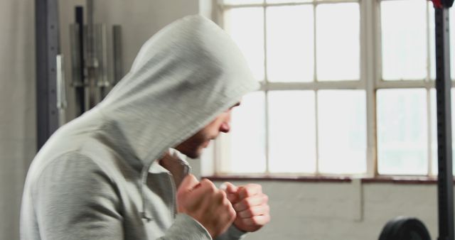 Man in grey hoodie shadowboxing near window in gym. Versatile image for fitness and workout related content. Suitable for articles or advertisements about physical training, exercise routines, and personal fitness journeys.