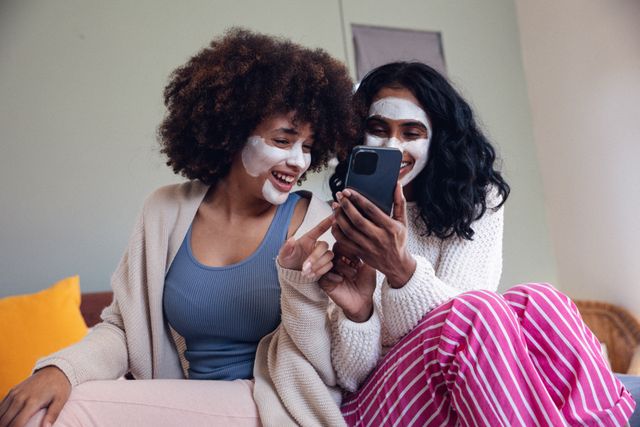Two happy biracial female friends with beauty creams on their faces are sitting on a bed and using a mobile phone. They are enjoying a casual and relaxing moment together, bonding over skincare and technology. This image is perfect for promoting beauty products, skincare routines, friendship, and leisure activities at home.