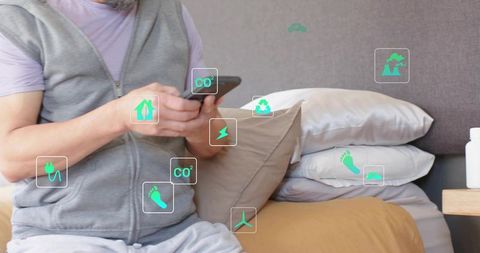Asian man tapping smartphone on bed, viewing green energy icons for smart home control